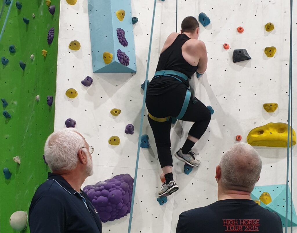 Danielle climbing a wall at indoor rock climbing supported by Jim and Richard.