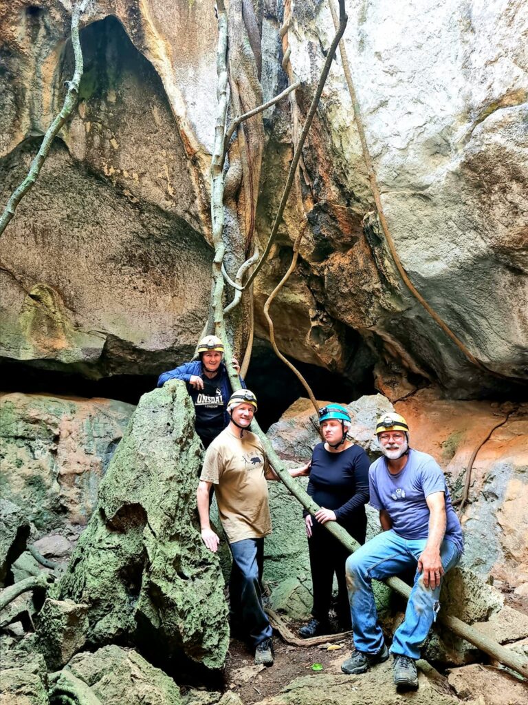 Adventure Image -  Lone, Richard, Danielle and Jim in an open section of cave. Large rock on the base of the cave and large fig tree roots climbing the walls of the cave.